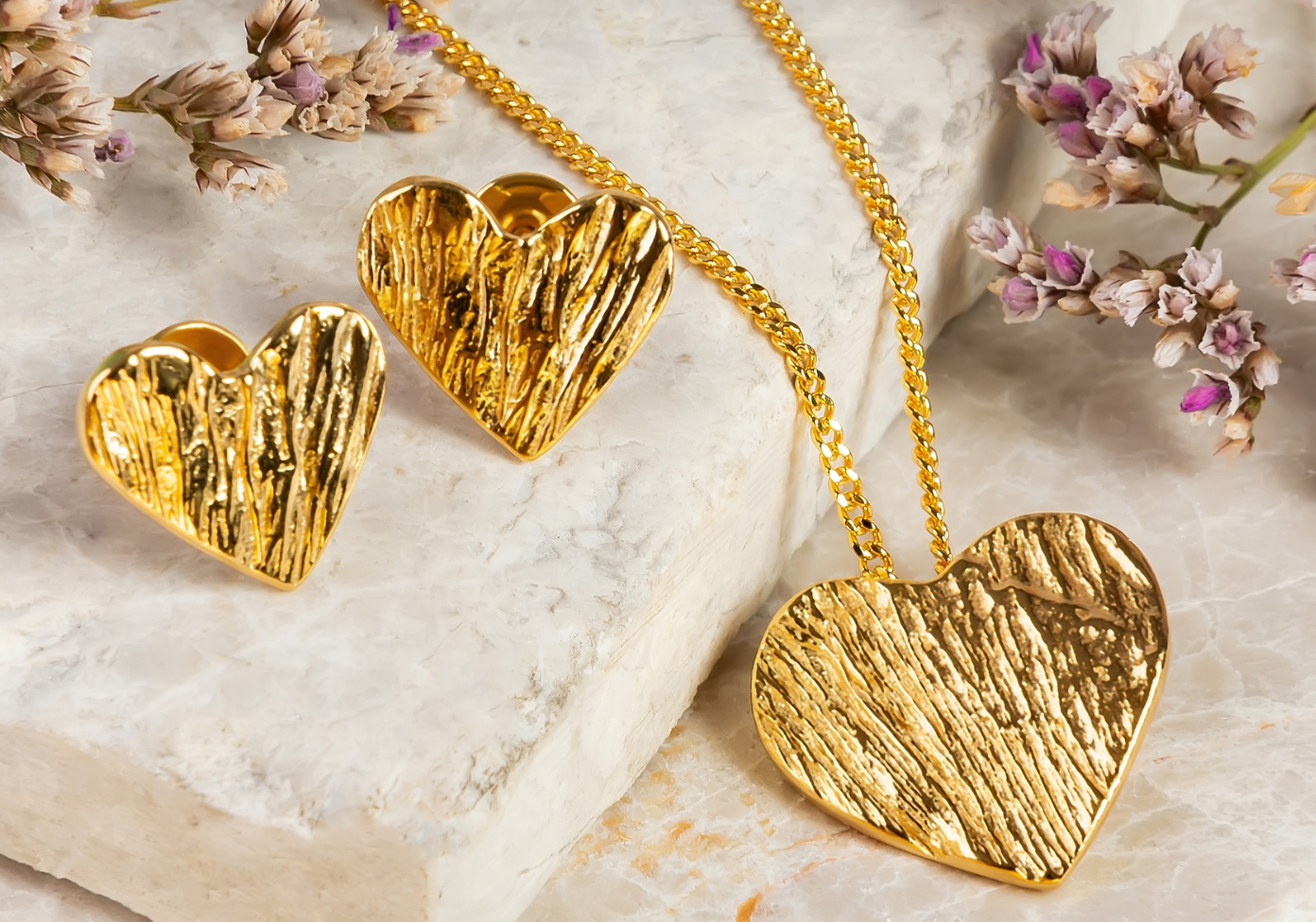 Gold heart-shaped jewelry set on a marble surface with dried flowers.